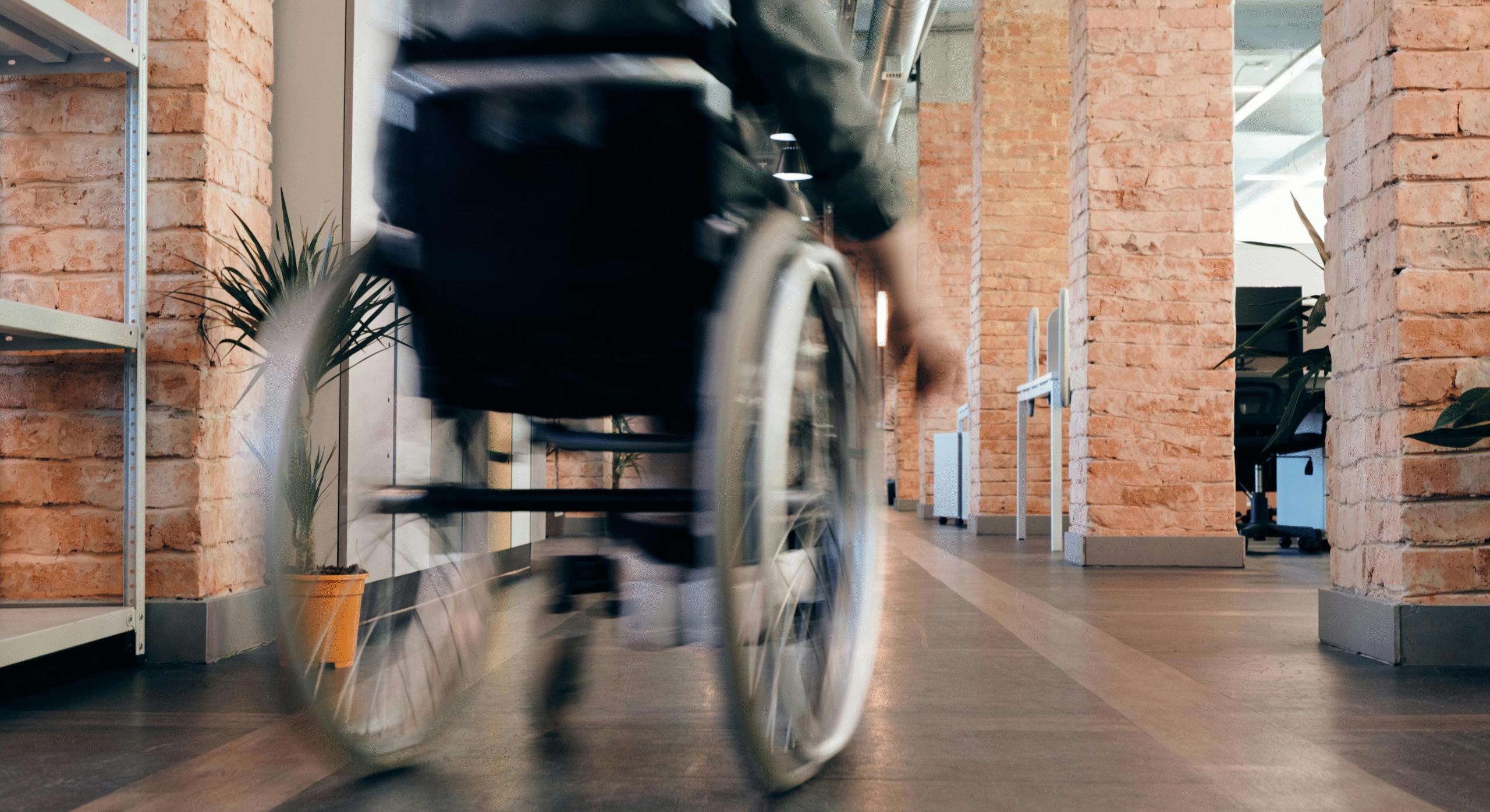 Eine Person im Rollstuhl bewegt sich anmutig durch einen Flur, der von Backsteinmauern und einer Topfpflanze gesäumt ist.